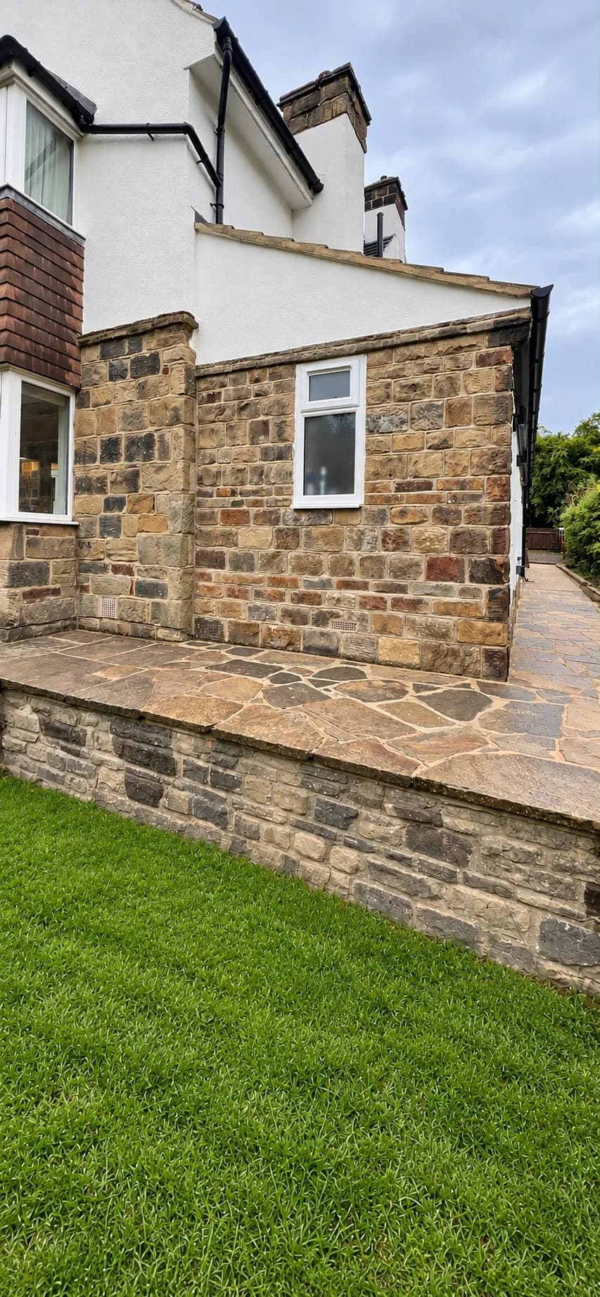 Stone walls and a white window along the side of the house, above a raised paved terrace and the lawn
